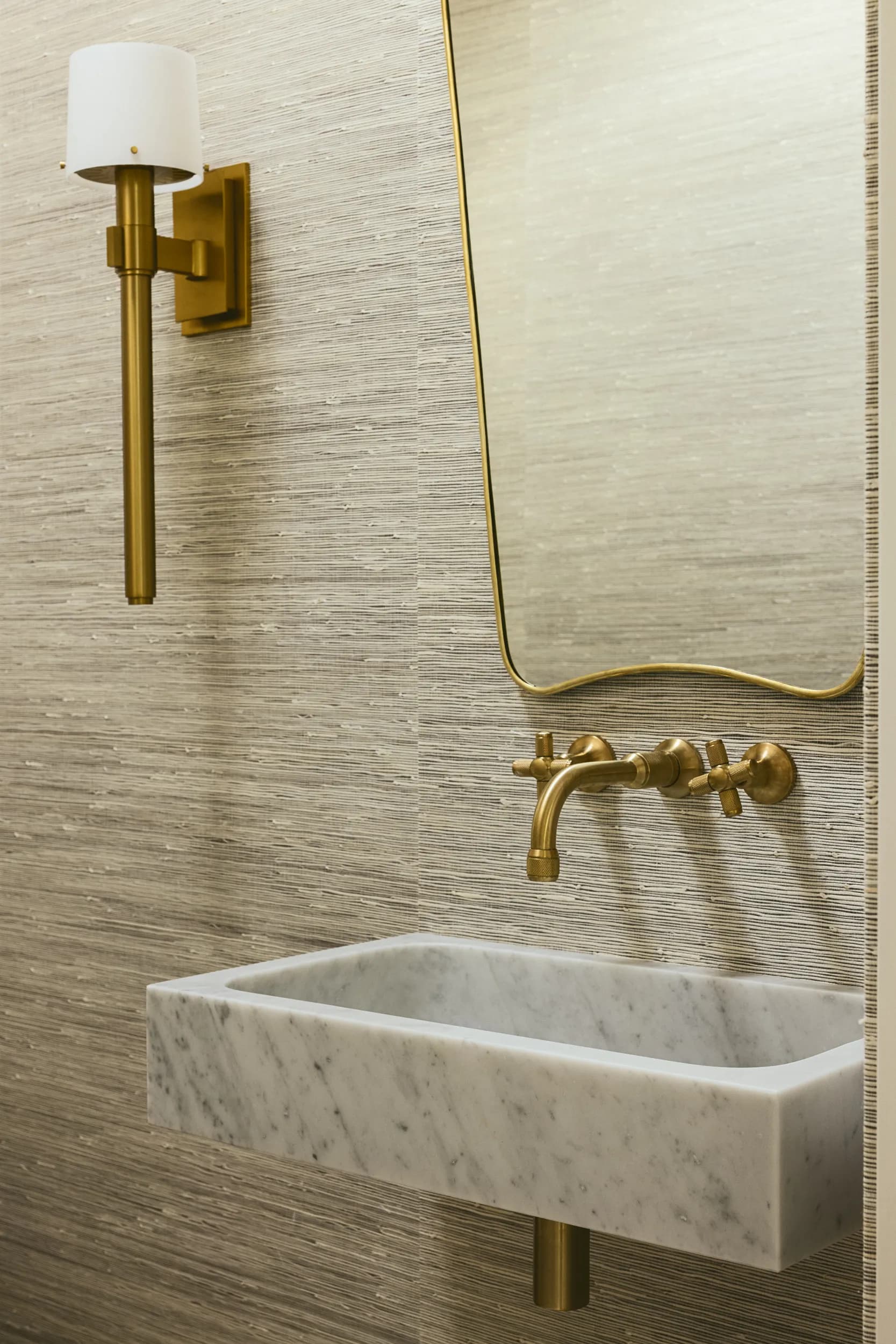 Elegant powder room featuring carved marble sink, brass wall-mount faucet, textured grasscloth wallpaper and brass sconce lighting House Rupert interior design Santa Barbara and Montecito