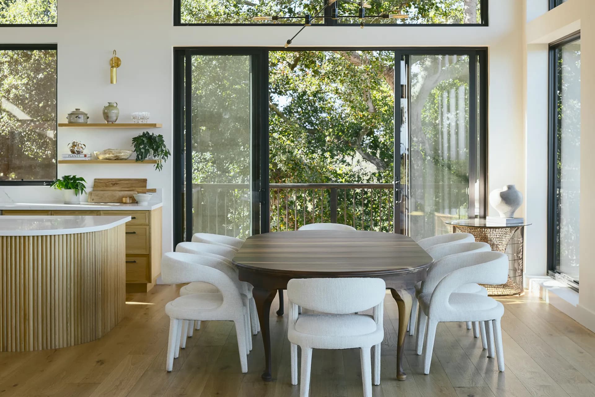 Modern dining room with dark wood round table, white upholstered chairs, fluted wood kitchen island, and large black-framed windows overlooking greenery, House Rupert interior design Santa Barbara and Montecito