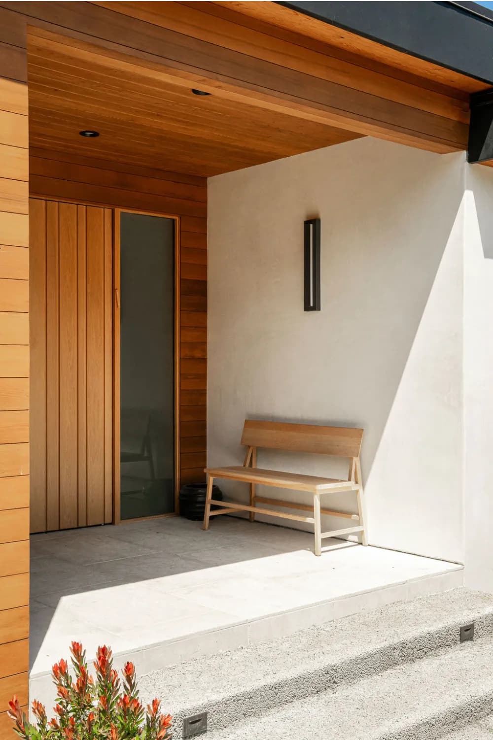 Modern covered entryway featuring warm cedar wood siding, glass door, minimalist wooden bench, and white stucco walls House Rupert interior design Santa Barbara and Montecito
