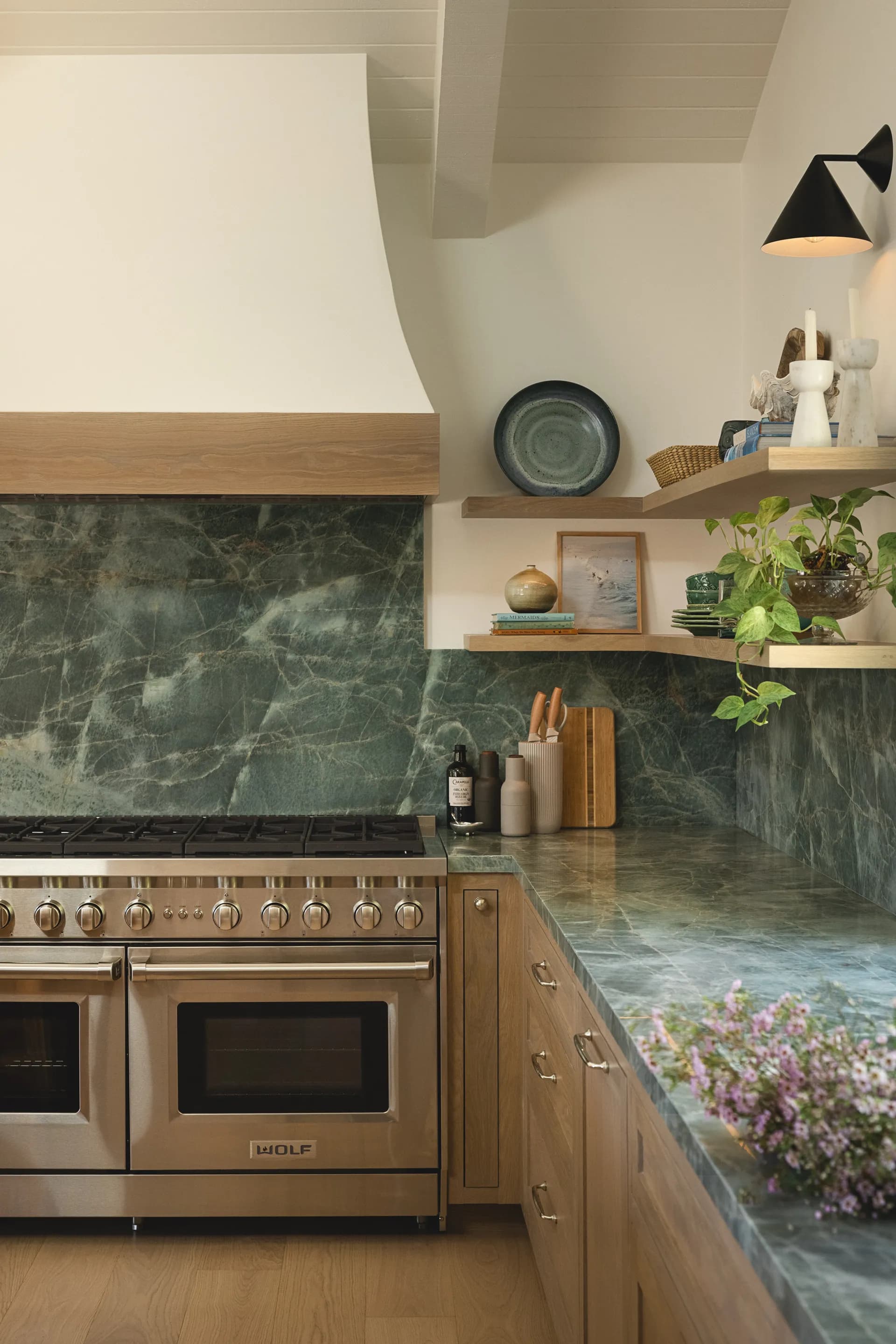 Modern kitchen featuring dramatic green marble backsplash, warm wood cabinetry, stainless steel Wolf range, and floating shelves with curated accessories House Rupert interior design Santa Barbara and Montecito