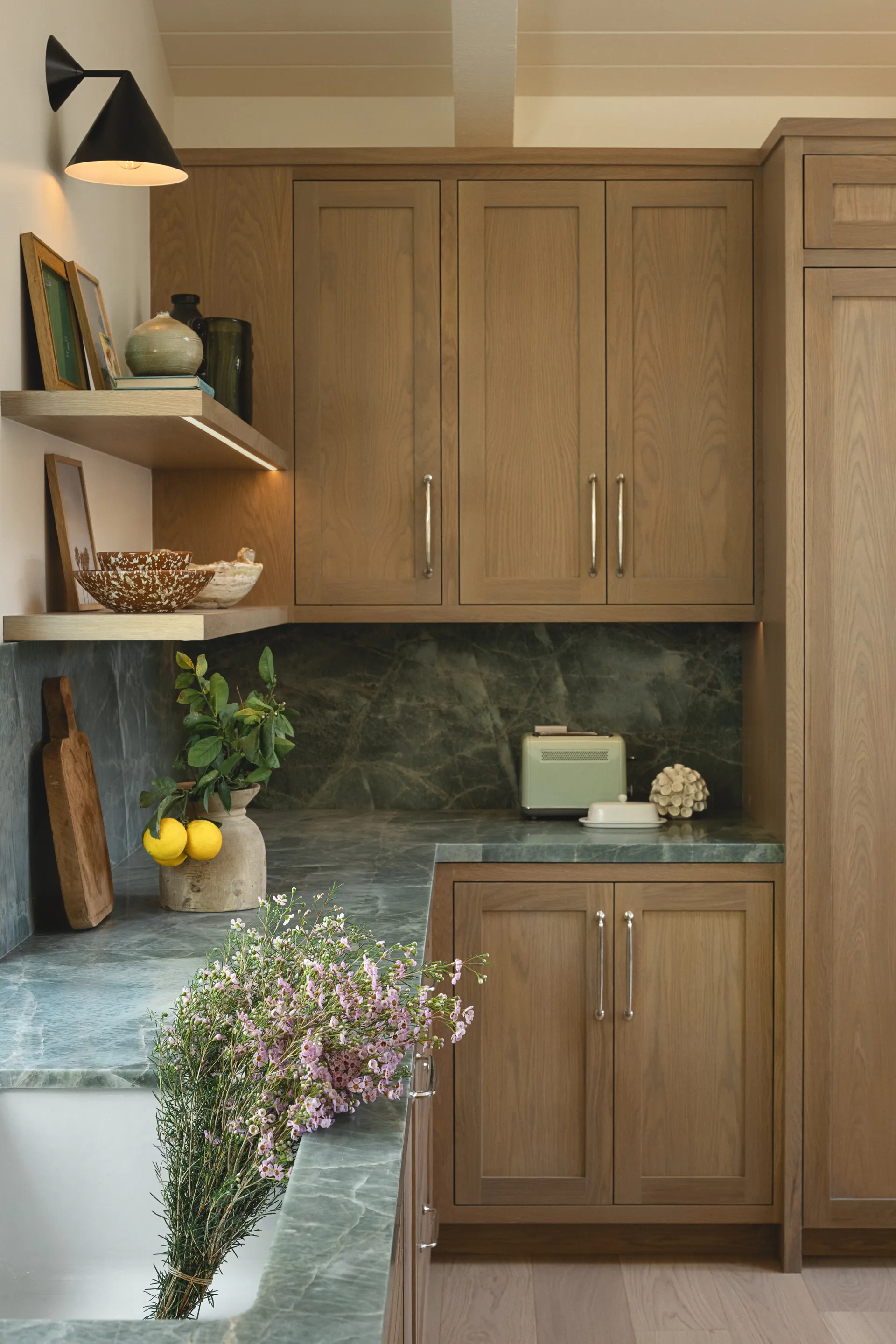 Warm oak kitchen cabinets with green marble countertops and backsplash, floating shelves, black sconce lighting, House Rupert interior design Santa Barbara and Montecito