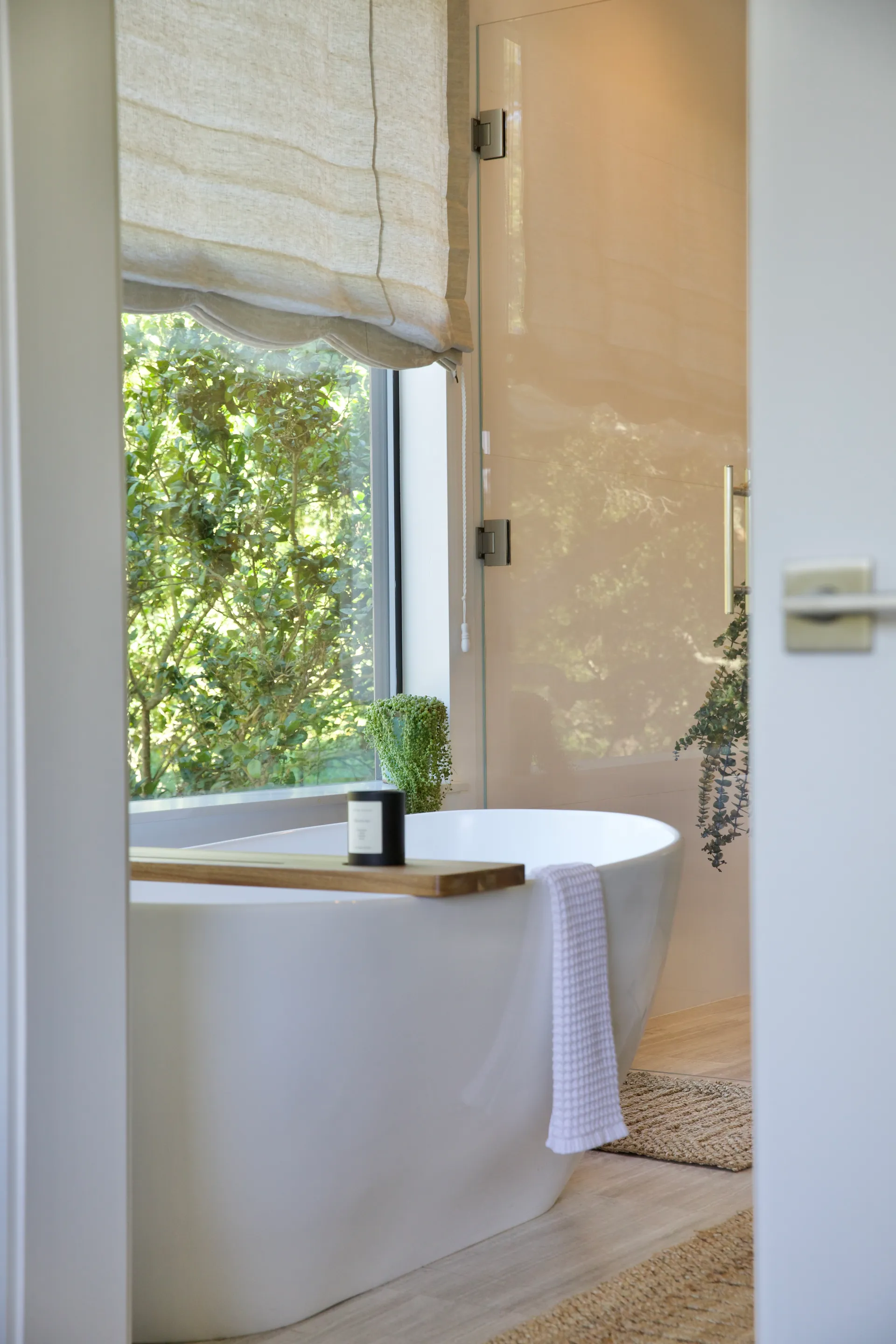 Modern bathroom featuring white freestanding bathtub with wooden caddy, roman shade window overlooking greenery, natural stone flooring House Rupert interior design Santa Barbara and Montecito