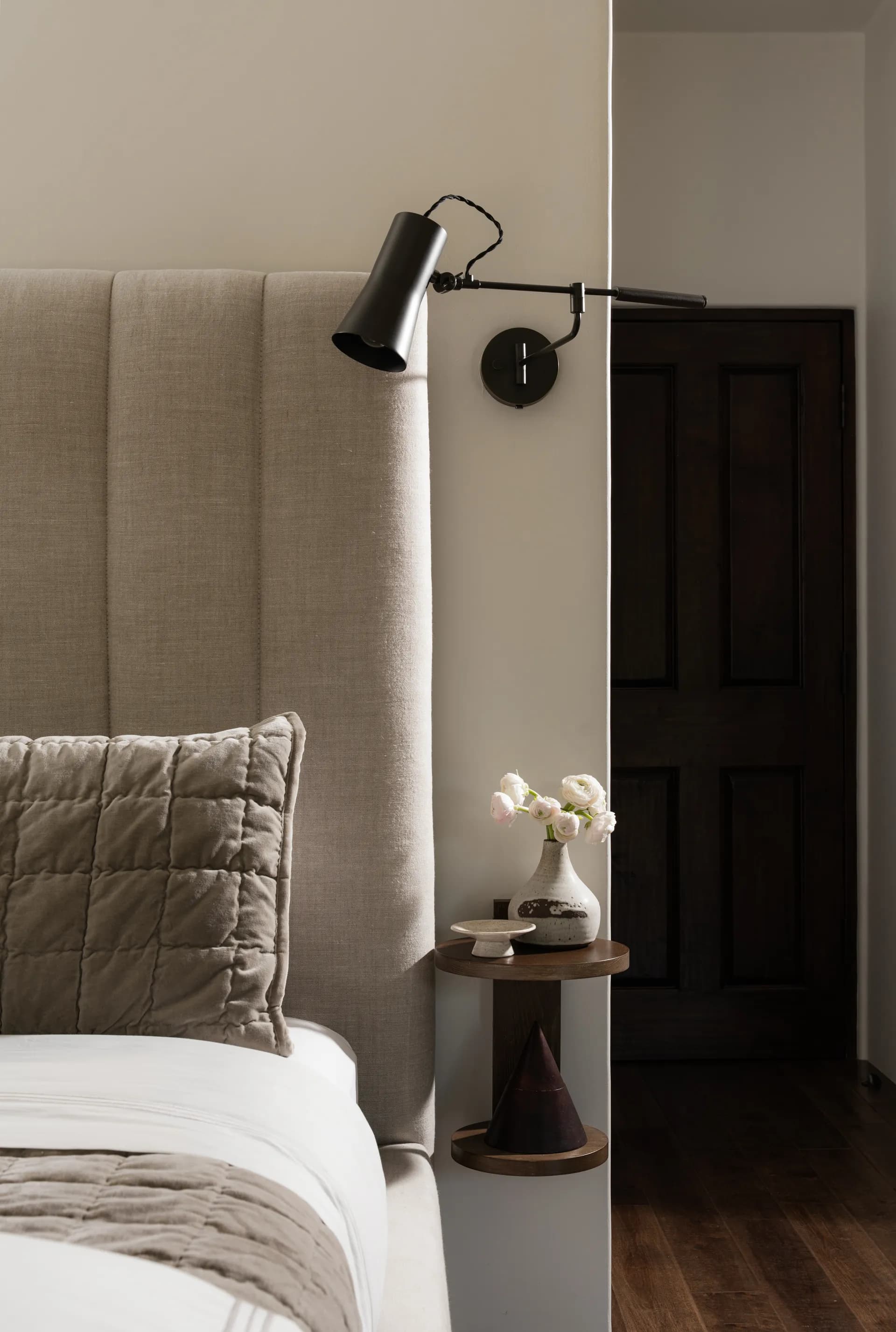 Modern bedroom featuring beige upholstered headboard, black adjustable wall sconce, quilted pillow, and round wooden nightstand with white ceramic vase House Rupert interior design Santa Barbara and Montecito