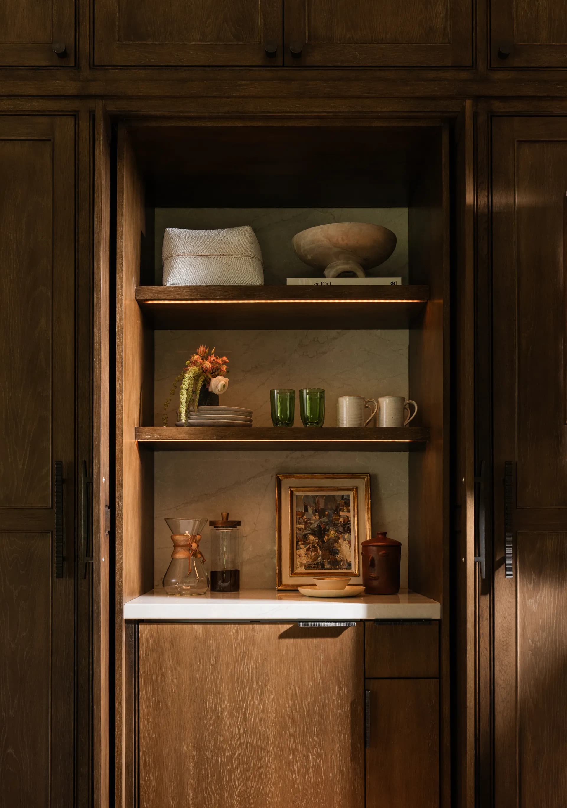 Custom walnut built-in shelving niche displaying ceramic vessels, green glassware, and coffee accessories with warm LED lighting House Rupert interior design Santa Barbara and Montecito