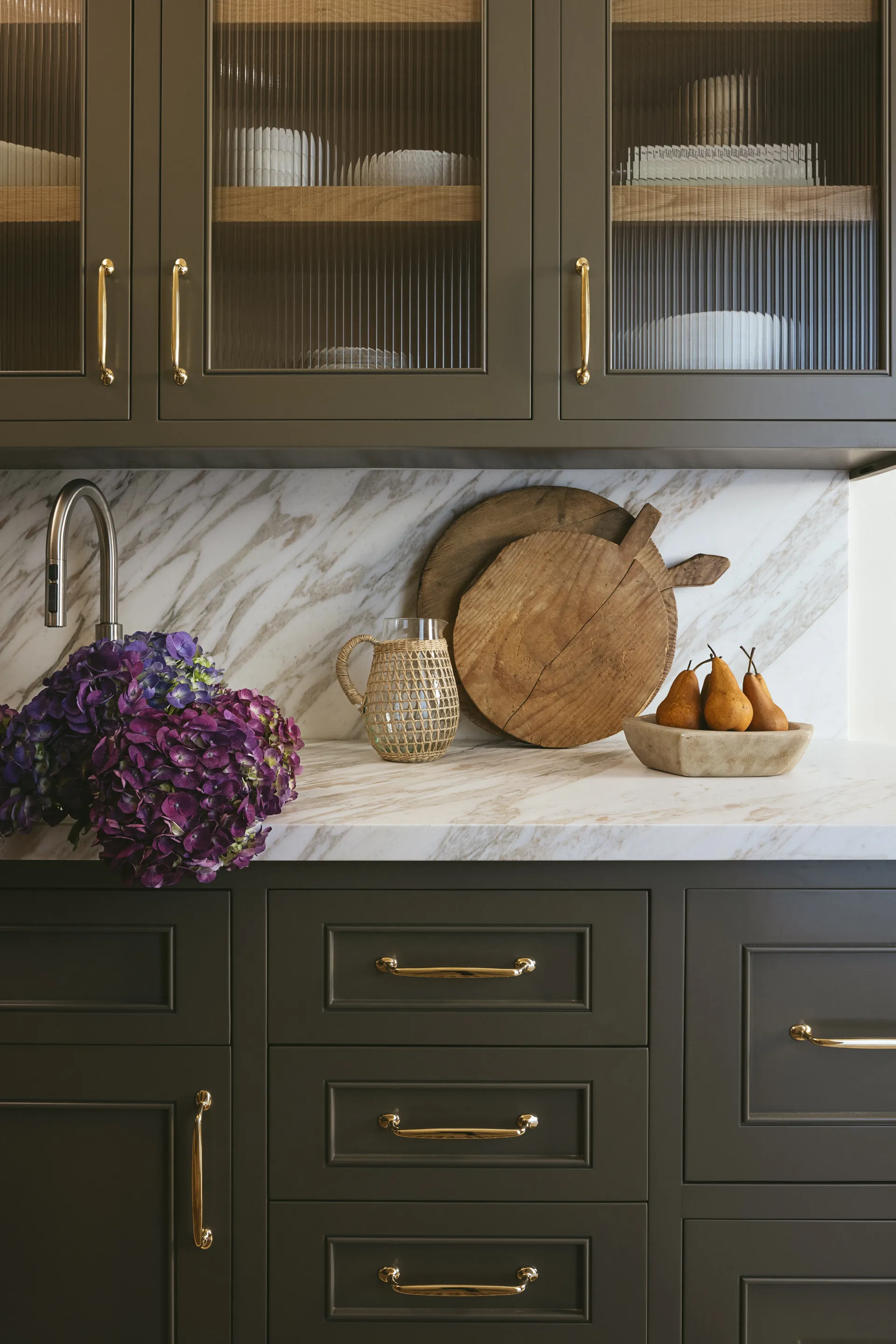 Dark green kitchen cabinets with fluted glass doors, Calacatta marble countertops, and brass hardware featuring purple hydrangeas and wooden cutting boards House Rupert interior design Santa Barbara and Montecito
