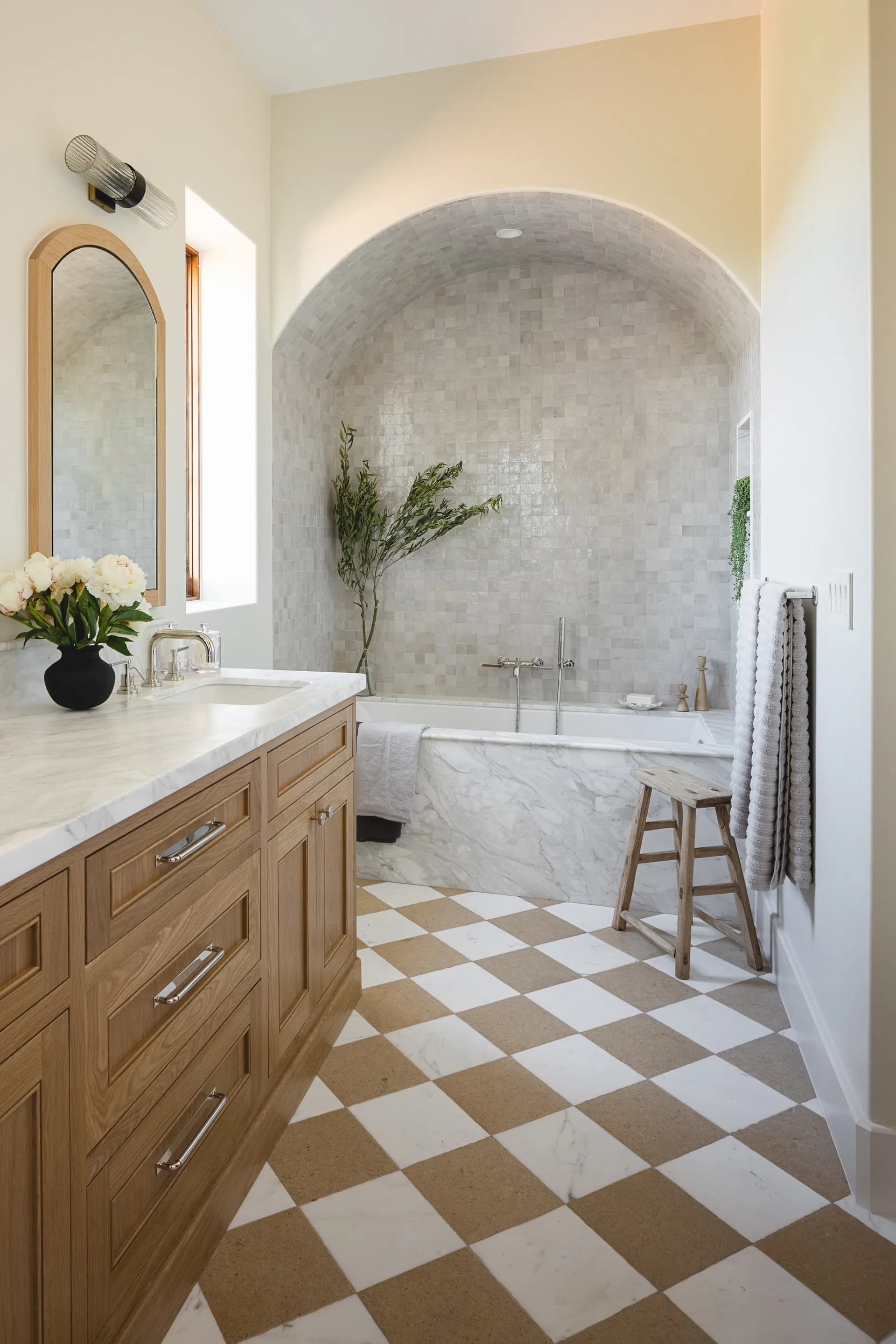 Mediterranean-style bathroom featuring arched marble tub niche, natural wood vanity, and geometric checkered stone flooring House Rupert interior design Santa Barbara and Montecito
