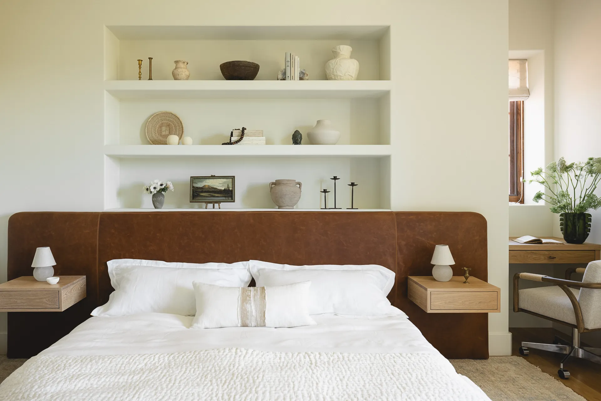 Contemporary bedroom featuring brown leather headboard, white built-in shelving with curated objects, floating nightstands and neutral textiles House Rupert interior design Santa Barbara and Montecito