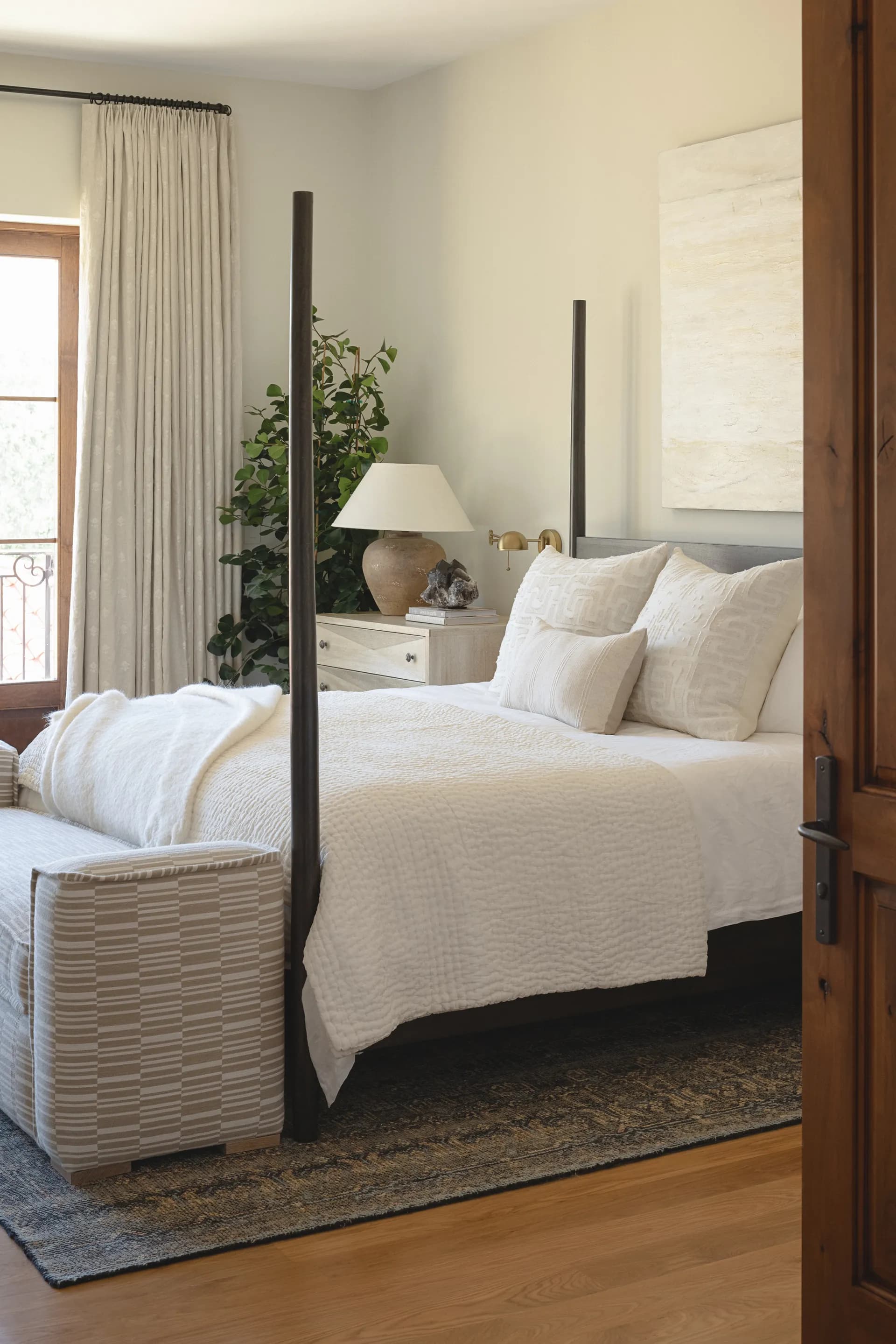 Serene bedroom featuring black four-poster bed with white linens, woven bench, linen curtains, and natural wood floors House Rupert interior design Santa Barbara and Montecito