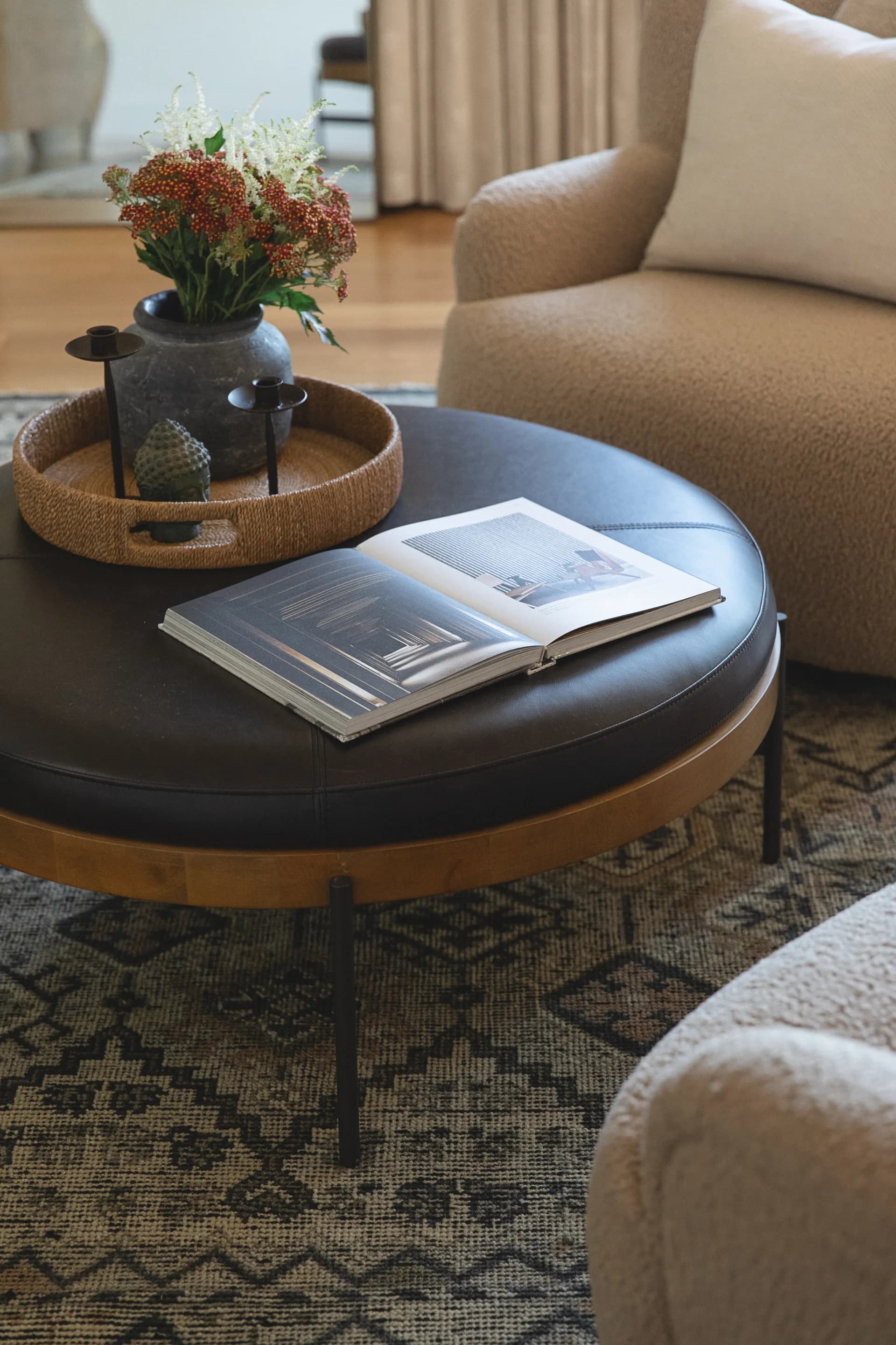 Round black coffee table with open book and woven tray holding vase with flowers beside beige upholstered chair House Rupert interior design Santa Barbara and Montecito