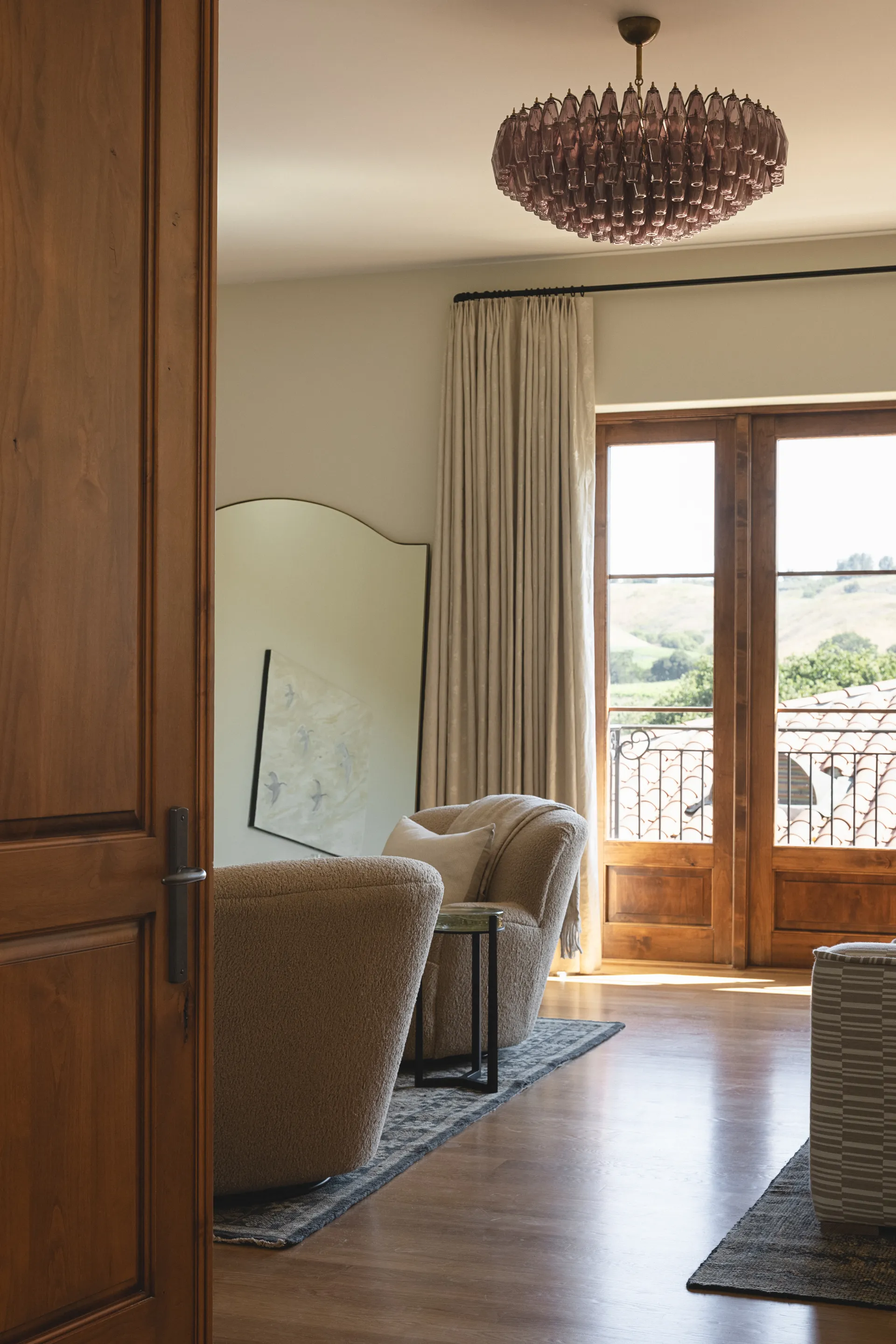 Warm living room featuring beige upholstered chairs, statement chandelier, wood paneling, and French doors opening to balcony, House Rupert interior design Santa Barbara and Montecito