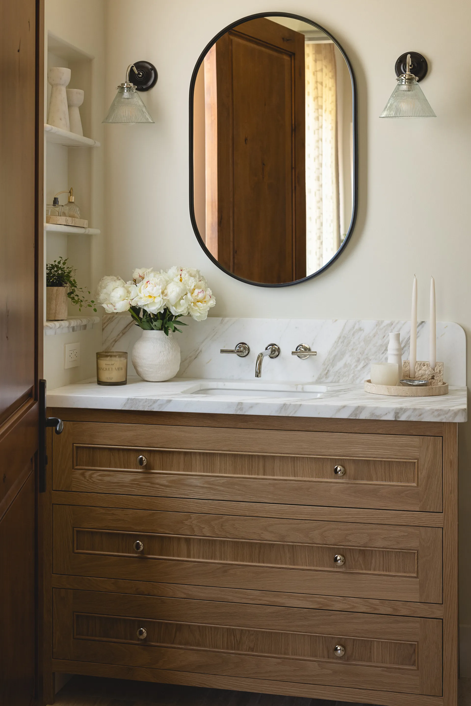 Warm wood bathroom vanity with oval black mirror, white marble countertop, wall-mount faucet, and glass sconce lighting House Rupert interior design Santa Barbara and Montecito