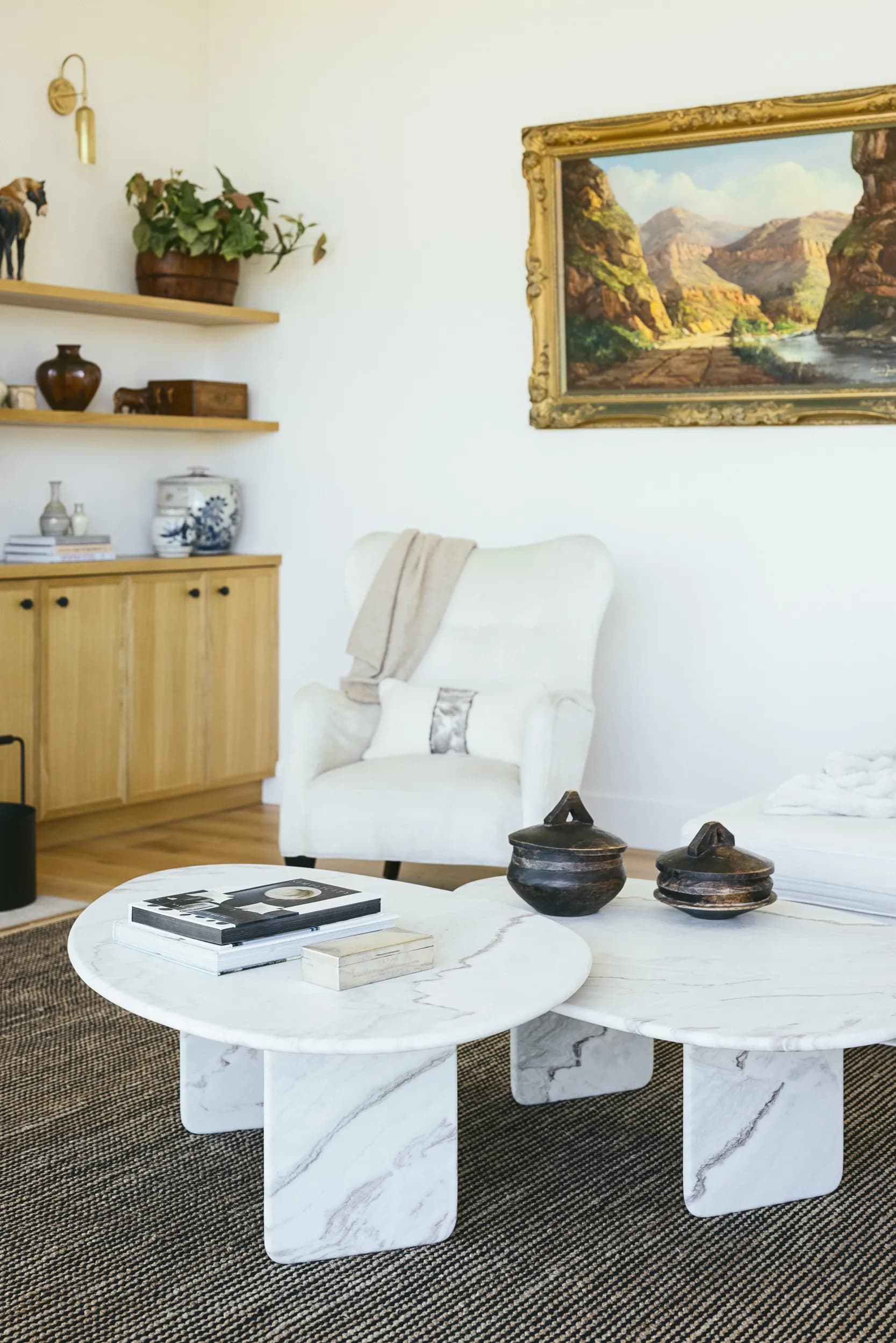 Round white marble coffee table with books and pottery beside cream upholstered chair beneath framed landscape painting House Rupert interior design Santa Barbara and Montecito
