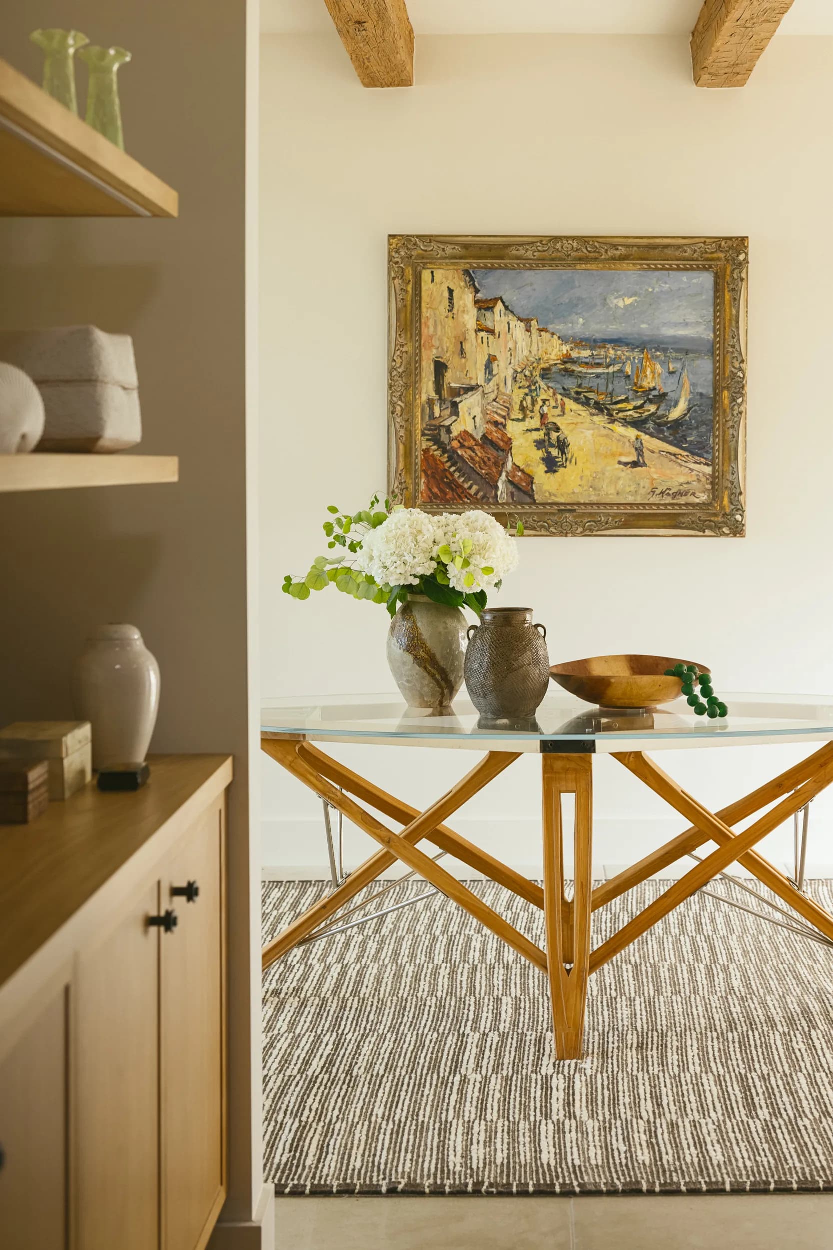 Mediterranean-style dining nook with glass-top table, wooden X-base, coastal painting, exposed ceiling beams, neutral tones House Rupert interior design Santa Barbara and Montecito