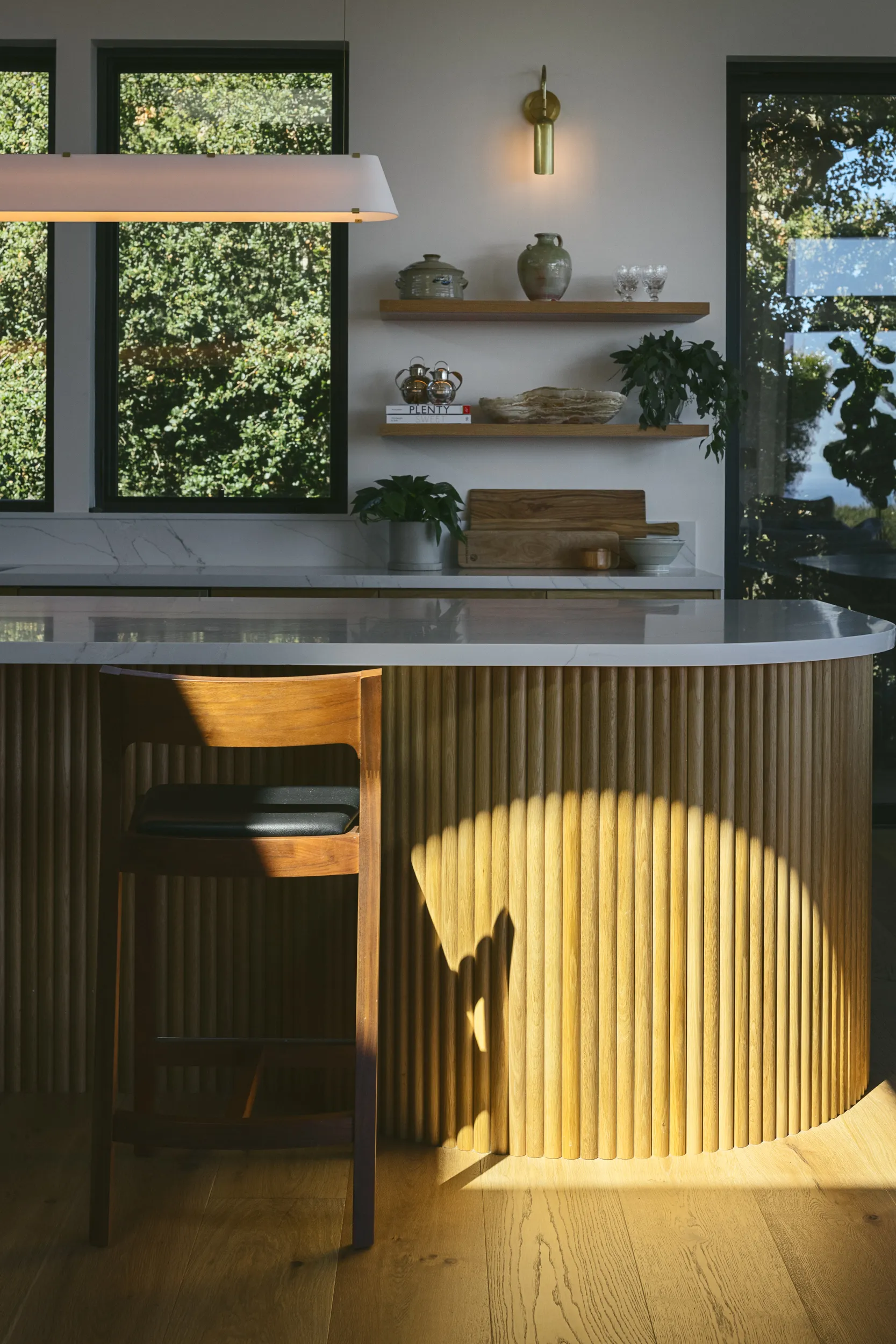 Modern kitchen island featuring fluted wood base with marble countertop, floating shelves, and brass sconce lighting House Rupert interior design Santa Barbara and Montecito