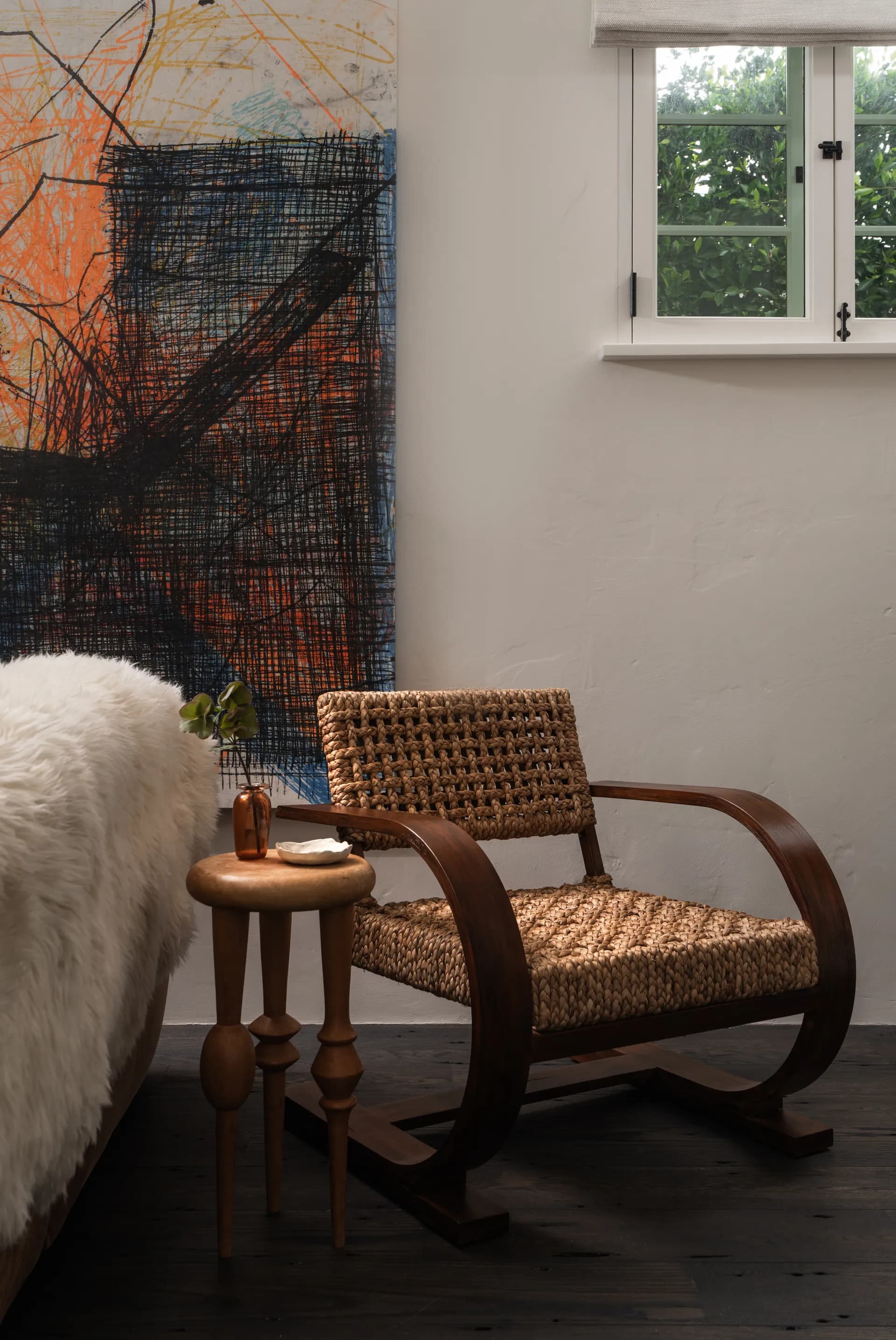 Woven rattan cantilever chair with curved wood frame beside abstract expressionist artwork and white fur throw, House Rupert interior design Santa Barbara and Montecito