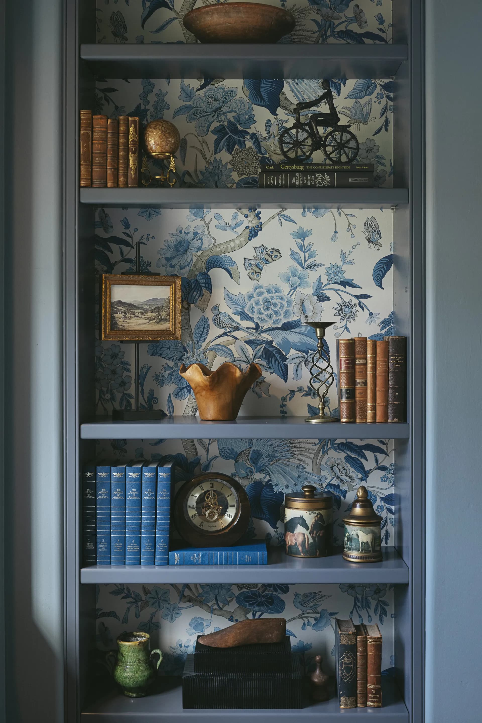 Built-in bookshelf with blue botanical wallpaper backdrop displaying vintage books, brass candlestick, ceramic vessels and antique collectibles House Rupert interior design Santa Barbara and Montecito