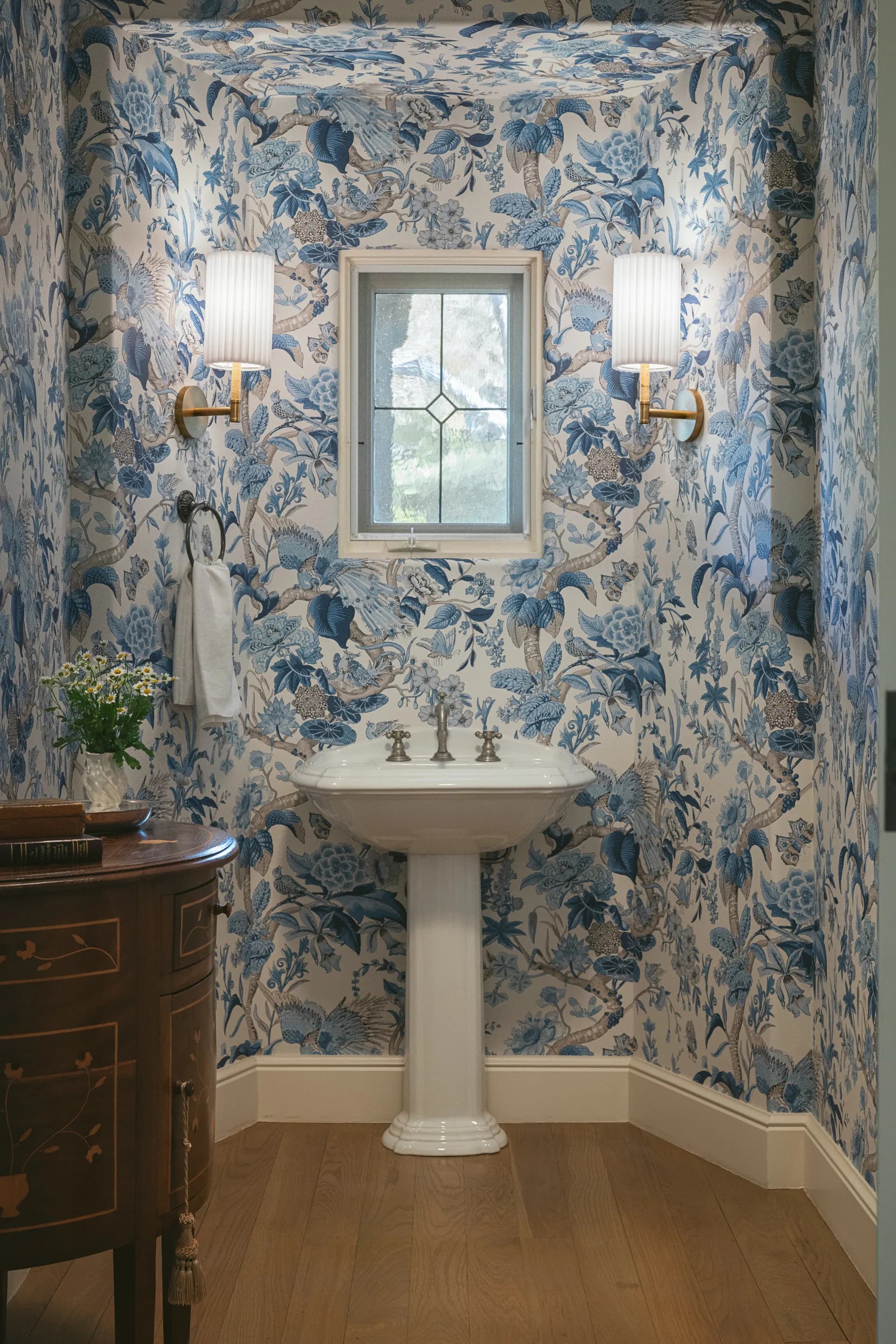Elegant powder room featuring blue and white floral wallpaper, white pedestal sink, brass sconces, and leaded glass window, House Rupert interior design Santa Barbara and Montecito
